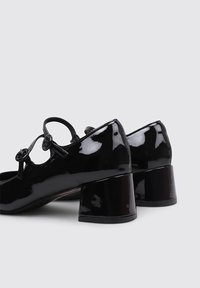 Chaussures en cuir verni noir avec un bout arrondi et un petit talon carré. Présente deux accents circulaires et une fine lanière sur le pied.