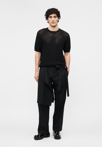 Άνδρας με σγουρά μαλλιά, που φοράει μαύρο πλεκτό τοπ με κοντά μανίκια και φαρδιά μαύρη παντελόνια με ασύμμετρο ντεκολτέ και μαύρα παπούτσια, που στέκεται μπροστά σε λευκό φόντο.