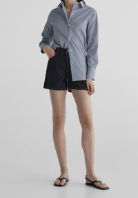 Marineblå och vit randig skjorta med knappar, kombinerad med högmidjade mörka denimshorts. Modellen bär svarta flip-flop-sandaler. Slät, avslappnad textur.