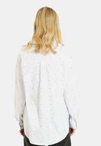 Camicia bianca con bottoni, caratterizzata da un motivo a pois luccicanti, maniche lunghe e orlo arrotondato, vista dal retro.