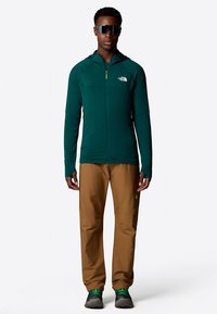 Zielona kurtka zapinana na zamek z żółtymi akcentami, brązowe spodnie i szare buty trekkingowe. Dodatki obejmują okulary przeciwsłoneczne. Materiał wydaje się gładki i lekki.