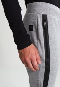 Graue Sweatpants mit schwarzen Seitenstreifen, Reißverschlusstasche und Stoffpatch-Label. Weiche Textur und lässiges Design.