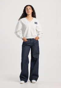 Light gray, V-neck sweatshirt with ribbed cuffs and a dark blue '89' print on the left chest, paired with wide-legged navy striped pants.