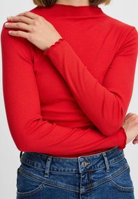 Rotes geripptes Langarmoberteil mit hohem Kragen und gewellten Handgelenkabschlüssen, kombiniert mit blauen Jeans aus Denim, die einen Knopf und Taschendetails zeigen.