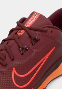 Zapatilla deportiva texturizada en color burdeos con acentos en naranja, suela de goma y cordones en tono similar. Presenta un logo en la lengüeta y una forma distintiva.
