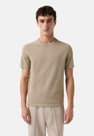 Beige pletený svetr s krátkým rukávem a kulatým výstřihem, se subtilní texturou, zkombinovaný se světlými, volně střiženými kalhotami.