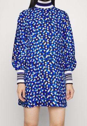 Jurk met lange mouwen in een patroon van blauwe, zwarte en witte golfjes, hoge gestreepte kraag en geribbelde gestreepte manchetten, gedragen door een model tegen een effen achtergrond.