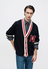 Μπλε πλεκτή ζακέτα με κόκκινες και λευκές λεπτομέρειες, με μεγάλο μπαλώμα με το γράμμα "H" και κλείσιμο με κουμπιά. Συνδυασμένη με καρό πουκάμισο και τζιν παντελόνι.