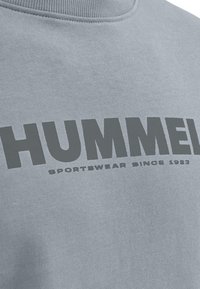 Sudadera azul claro con cuello redondo, que presenta un gran texto gris "HUMMEL" y "SPORTSWEAR SINCE 1923" en una fuente en negrita. Textura de tela suave.