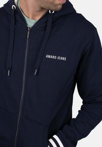 Sudadera con capucha azul marina con cremallera, fabricada con tejido suave, con el logo blanco "AMARO JEANS", capucha con cordón y puños acanalados con rayas.