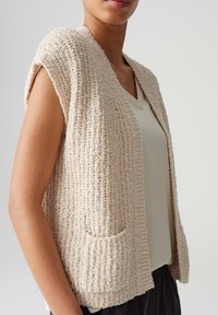 Beige gestrickte Weste mit strukturiertem Oberflächenmaterial, offener Front, ohne Ärmel und zwei Vordertaschen. Übereinander getragen mit einem hellen V-Ausschnitt Oberteil.