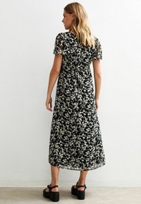 Mujer con cabello rubio que lleva un vestido midi negro con estampado floral blanco y amarillo, mangas cortas abullonadas y sandalias de plataforma negras, de pie.