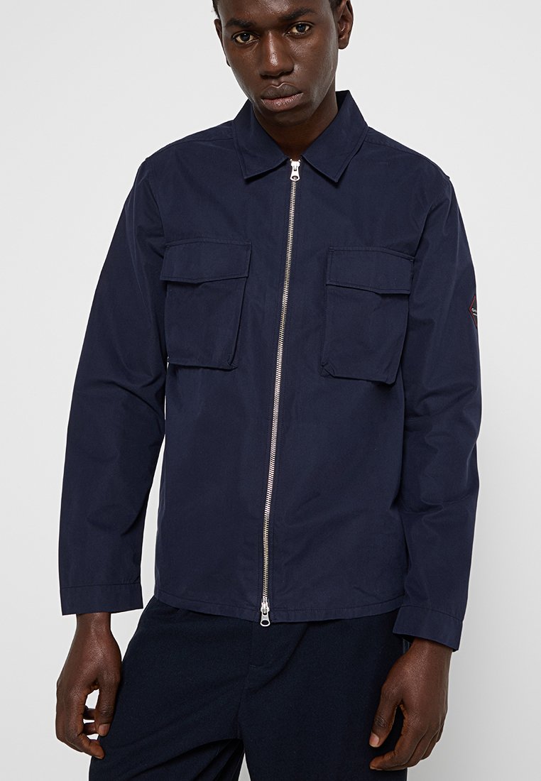 Chaqueta azul marino con cierre frontal, dos bolsillos en el pecho y un cuello. Fabricada con una tela ligera y suave con una textura sutil.