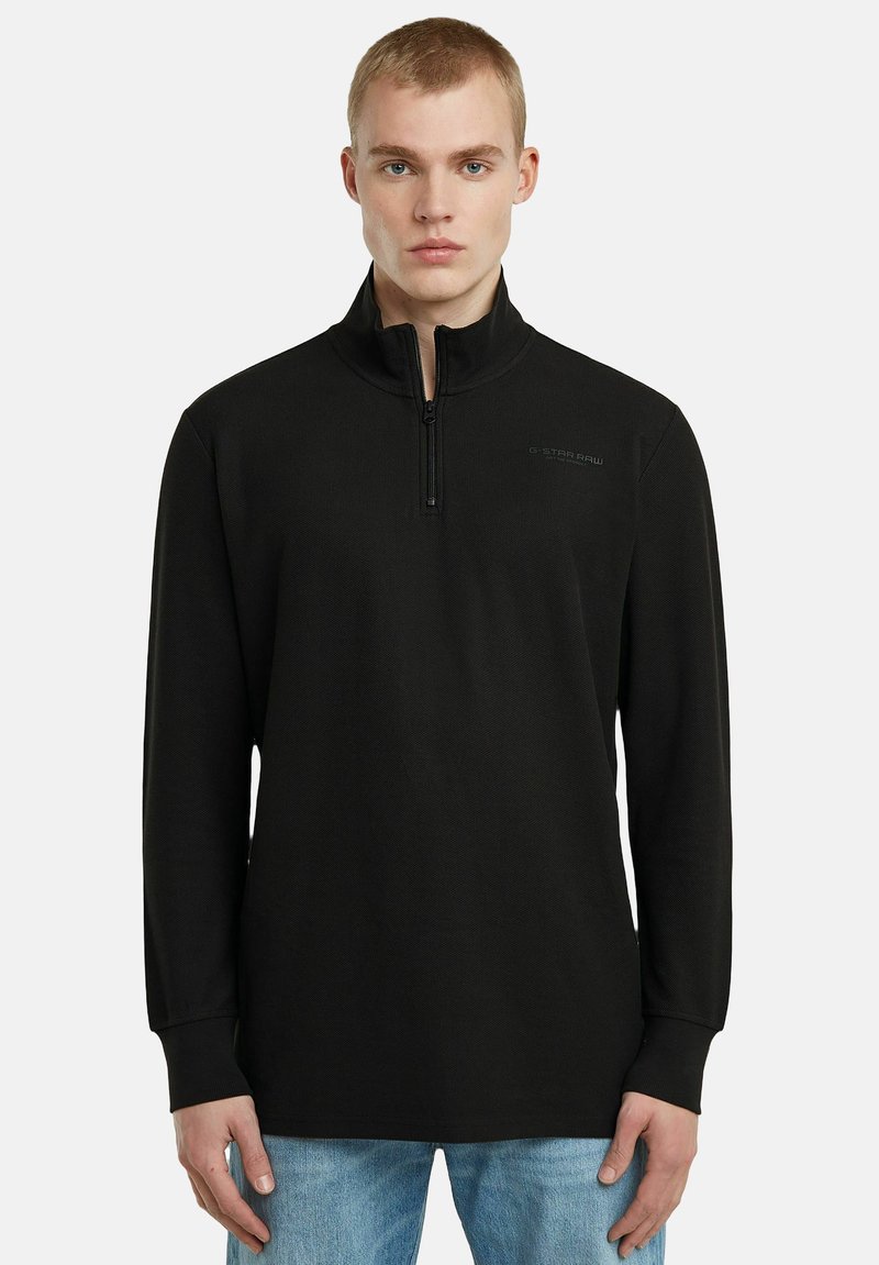 Schwarzer Pullover mit Viertelreißverschluss, langen Ärmeln, strukturiertem Stoff, mit einem kleinen Logo auf der linken Seite, kombiniert mit blauen Jeans.