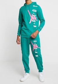 Man i teal Jordan-hoodie och matchande joggers med stor rosa Jumpman-logotyp och vita sneakers, stående mot en enkel bakgrund.