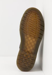 Semelle en caoutchouc affichant un motif texturé avec des rainures et des crêtes, de couleur brune, avec un logo circulaire en relief au centre.