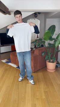 Una camiseta de algodón blanca sostenida por una persona, combinada con unos jeans azules y zapatillas plateadas, con un fondo de plantas de interior y muebles.