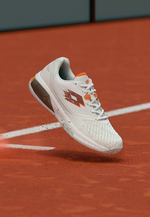 Weißer und oranger Sportschuh wirbelt Tonstaub auf einem roten Sand-Tennisplatz nahe einer weißen Begrenzungslinie auf.