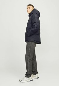 Marineblå pufferjakke med hætte, der har et quiltet design og sidelommer, båret over mørke jeans og lyse sneakers.