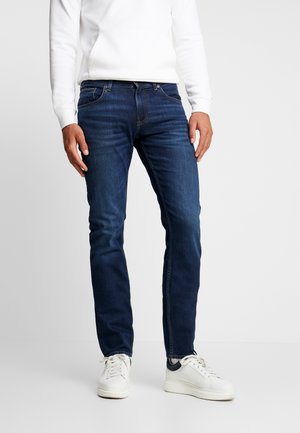 Mężczyzna w ciemnoniebieskich obcisłych dżinsach, białej koszuli z długim rękawem i białych tenisówkach stojący na tle jasnego, jednolitego tła.