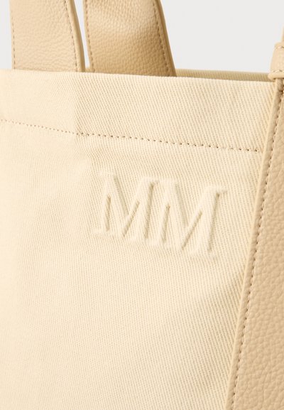 Κοντινό πλάνο μπεζ τσάντας tote από καμβά με αντιστοιχισμένες λαβές από δέρμα με υφή και ανάγλυφα γράμματα "MM" στην μπροστινή όψη.