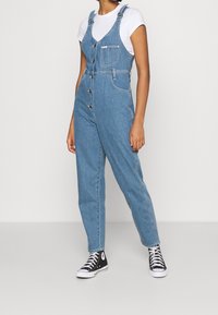 Blå denimoverall med en framficka, metallknappar och smalnade ben, i kombination med en vit t-shirt och svarta hög-top sneakers.