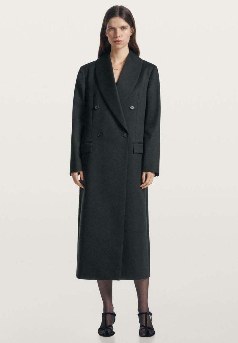 Dvouradý tmavě šedý vlněný kabát se širokými klopami, bočními kapsami a přizpůsobeným střihem, sahající do poloviny lýtek. Silueta je strukturovaná.