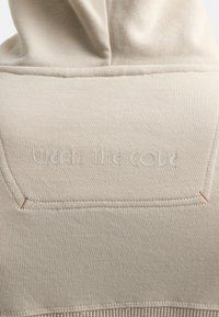 Beige hoodie med præget tekst "WEAR THE CODE" på ryggen. Stofstrukturen er blød, med synlige stikninger og et enkelt design.
