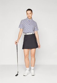 Koszulka golfowa z krótkim rękawem i kołnierzykiem, z niebiesko-białym wzorem tkanym, w połączeniu z czarną spódnicą i białymi trampkami. W ręku kij golfowy.