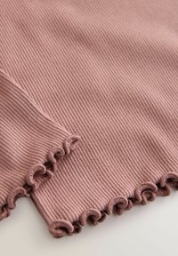 Prim-plan al unui material nervurat mov pal, cu margini volanate, pe o suprafață moale și texturată a unei haine.