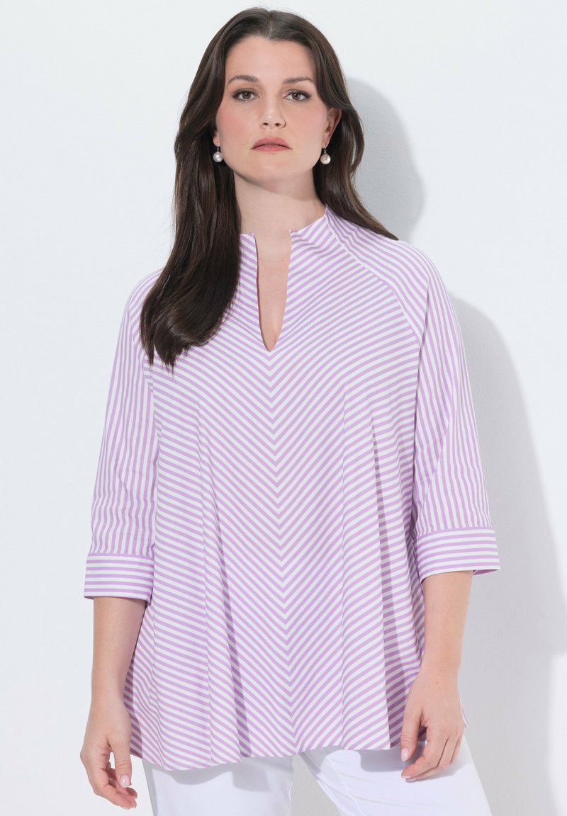 Donna con capelli lunghi e scuri che indossa orecchini di perle e una maglia a righe lavanda e bianca con maniche a tre quarti, in piedi contro un muro bianco.