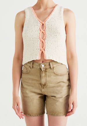 Femme portant un crop top blanc en maille sans manches avec bordure pêche et un short en jean beige taille haute sur un fond uni.