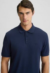 Poloko navy modrej farby z hladkej tkaniny, s golierom, trojbodovým zapínaním a jemným logom na hrudi. Krátke rukávy.