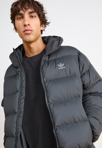 Sötétszürke puffertakaró, kapucnival, cipzáras záródással és steppelt textúrával. Az elülső részen Adidas logó található, fekete póló felett viselve.