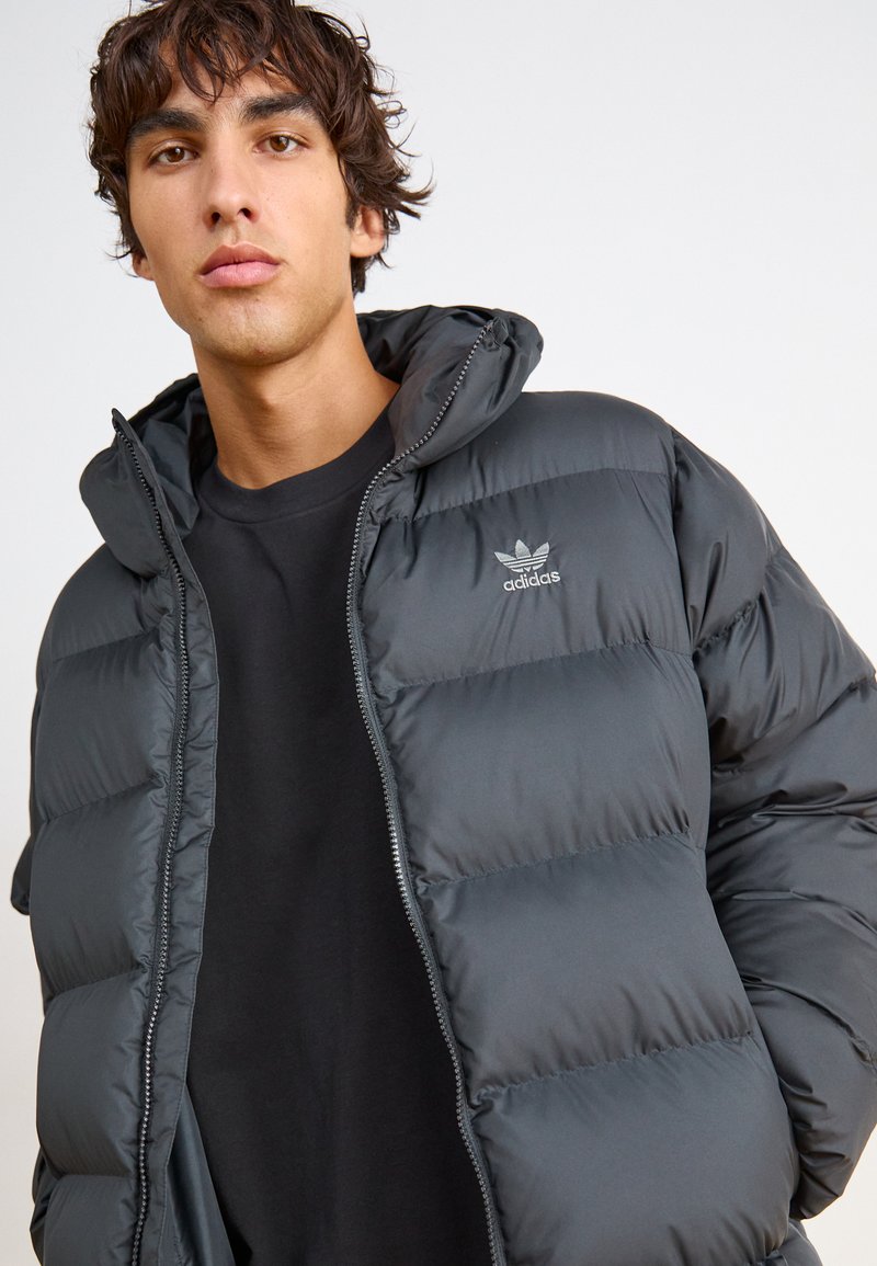 Dunkelgraue Pufferjacke mit Kapuze, Reißverschluss und gestepptem Muster. Mit einem Adidas-Logo auf der Brust, getragen über einem schwarzen Shirt.