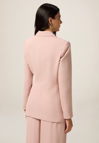 Lichtroze op maat gemaakte blazer van gladde stof, met een klassieke kraag, lange mouwen en een licht gebogen zoom. Achteraan zijn de strakke lijnen te zien.