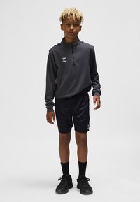 Sportoutfit bestaande uit een grijs lang overhemd met rits en zwarte korte broek. Het shirt heeft een gestructureerde afwerking; de korte broek heeft een glad oppervlak. Zwarte sneakers maken de look compleet.