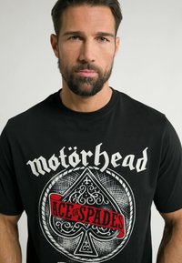 Sort bomulds T-shirt med et grafisk design, der har teksten "Motörhead" og "Ace of Spades" i rødt på et spar-emblem.