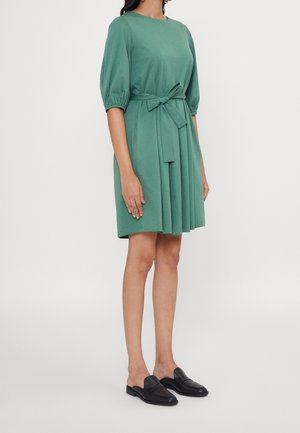 Femme portant une robe verte longueur genoux avec des manches bouffantes, une ceinture nouée à la taille et des chaussures noires à enfiler, se tenant devant un fond blanc.
