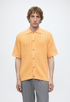 Jeune homme portant une chemise en maille à manches courtes couleur pêche avec des boutons et un pantalon gris, debout devant un fond blanc uni.