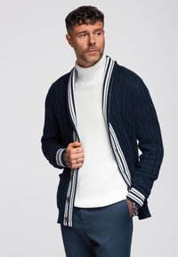 Cardigan in maglione a trecce blu navy con colletto a scialle a righe bianche, apertura frontale e dettagli con bottoni di legno. Indossato sopra un dolcevita bianco.