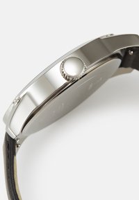 Edelstahl-Uhr mit silbernem Gehäuse, rundem Design und strukturiertem schwarzen Lederarmband. Sie verfügt über eine auffällige Krone an der Seite.