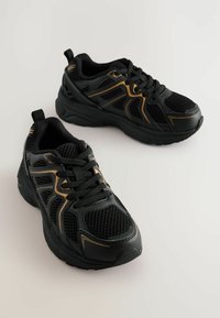 Chaussures de sport noires avec des matériaux en maille et synthétiques. Présentent des accents dorés, un système de laçage, et une semelle texturée. Inclut une languette à l'arrière.