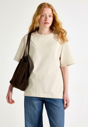 Ung kvinde med langt rødt hår iført en løs beige t-shirt, blå jeans og bærer en mørkebrun skuldertaske.
