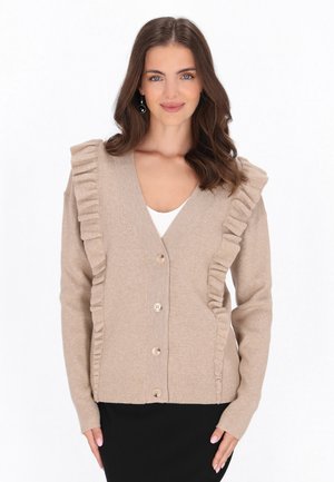 Frau mit schulterlangem braunem Haar, die eine beige, geknöpfte Strickjacke mit Rüschen und schwarzen Rock trägt und in die Kamera vor weißem Hintergrund lächelt.