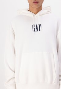 Hoodie blanc avec une poche avant, capuche à cordon de serrage et logo "GAP" bleu marine. Tissu doux avec des poignets et un ourlet inférieur côtelés.