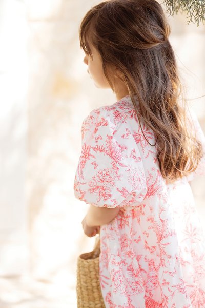 Vestido floral branco com padrões rosa, mangas bufantes, tecido leve e uma cesta de vime. Cabelo solto em ondas suaves.