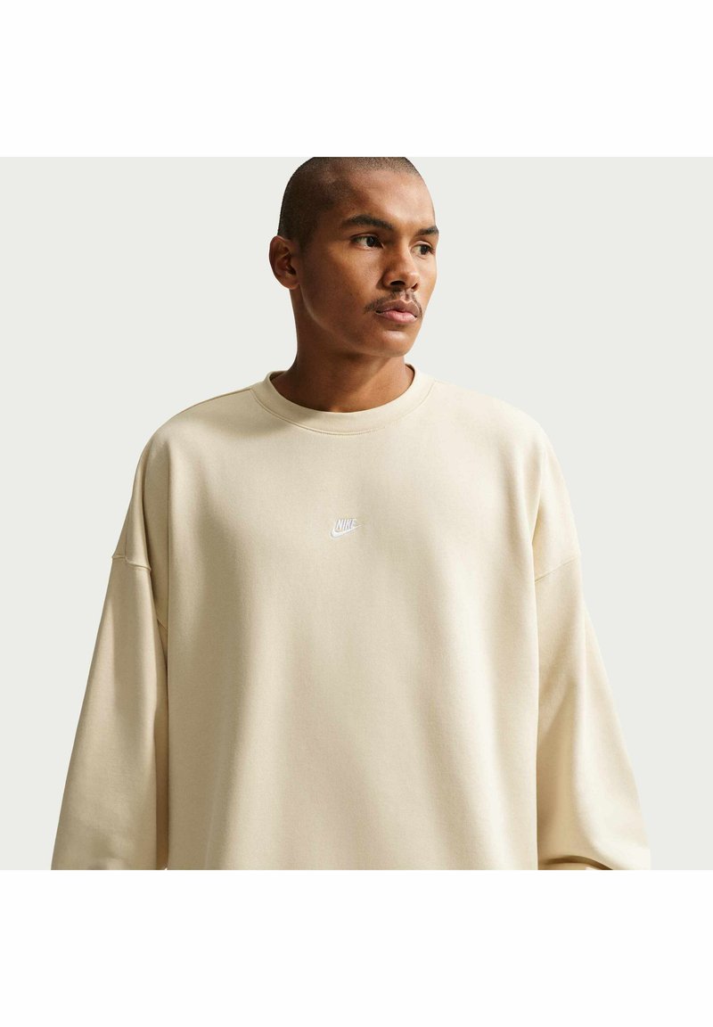 Homme aux cheveux courts portant un sweatshirt Nike beige oversize, regardant sur le côté devant un fond clair uniforme.