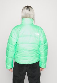 Person med lyst blondt hår iført en klar grøn pusletaske med The North Face-logoet, set bagfra mod en hvid baggrund.