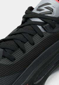 Μαύρο αθλητικό παπούτσι με υφή στην επιφάνεια, γκρι λεπτομέρειες και μαύρα κορδόνια. Διαθέτει γυαλιστερή μαύρη σόλα και χαρακτηριστική ετικέτα με λογότυπο κοντά στη γλώσσα.